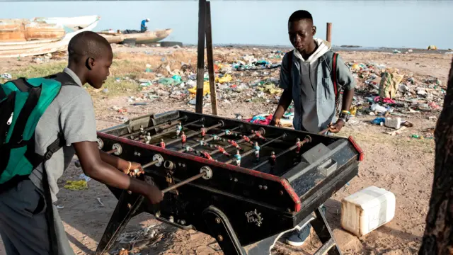 Deux écoliers jouent une partie de baby-foot sur les rives du fleuve Casamance, avec derrière eux des détritus et un homme qui s'occupe d'un bateau.