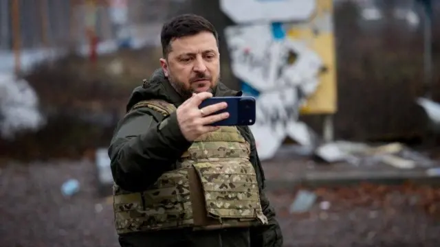 Владимир Зеленский держит телефон на фоне разрушенного Купянска