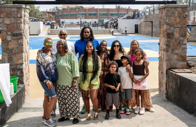 Kawhi Leonard wambaye umupira w'ubururu uhagaze inyuma hamwe n'abantu bo mu muryango we bari imbere y'ibibuga bya basketball ku ishuri rya St Ignace i Kibagabaga mu mujyi wa Kigali