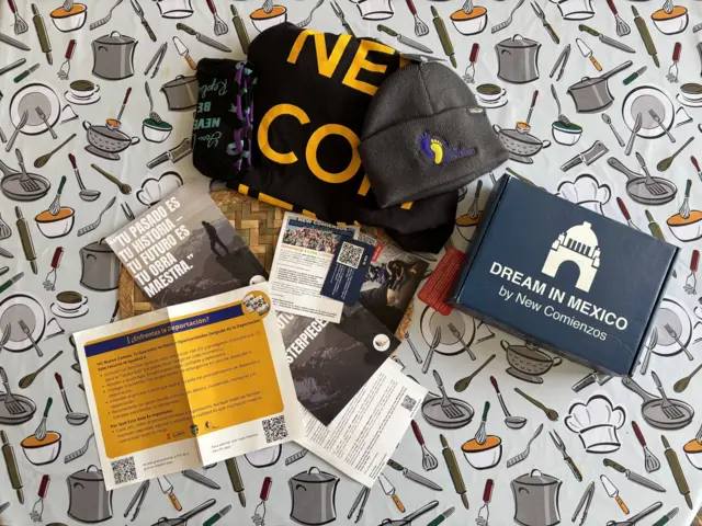 Artículos promocionales y folletos de New Comienzos, incluidos gorros, una bufanda y un libro, colocados sobre un mantel con ilustraciones de utensilios de cocina.