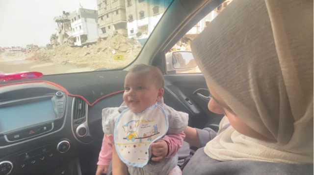 A six-month old baby smiles and is held up by her mother in the front seat of a car, against the backdrop of the destruction of southern Gaza visible from the front screen car window.