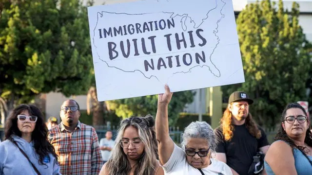 Manifestantes protestam contra uma deportação realizada pelo ICE em Los Angeles, na Califórnia (EUA). Uma manifestante, com a cabeça baixa, exibe um cartaz com os dizeres 'A imigração construiu esta nação'.