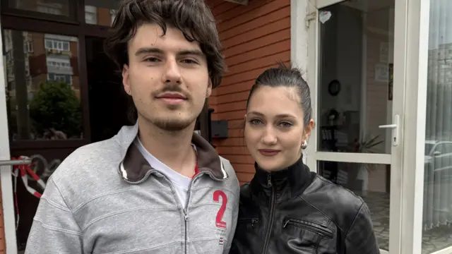 Адріан, одягнений у білу футболку та сіру куртку, стоїть поруч з Інгрід, одягненою в чорну шкіряну куртку, біля входу в багатоквартирний будинок.