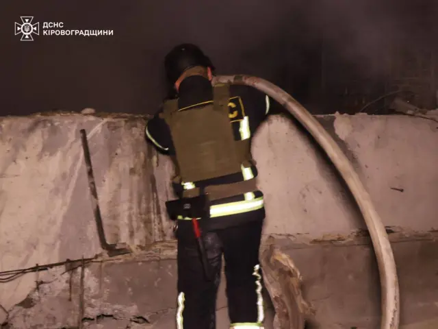 Пожежник спиною до камери біля зруйнованої стіни. Він тримає пожежний рукав і спрямовує воду кудись вниз