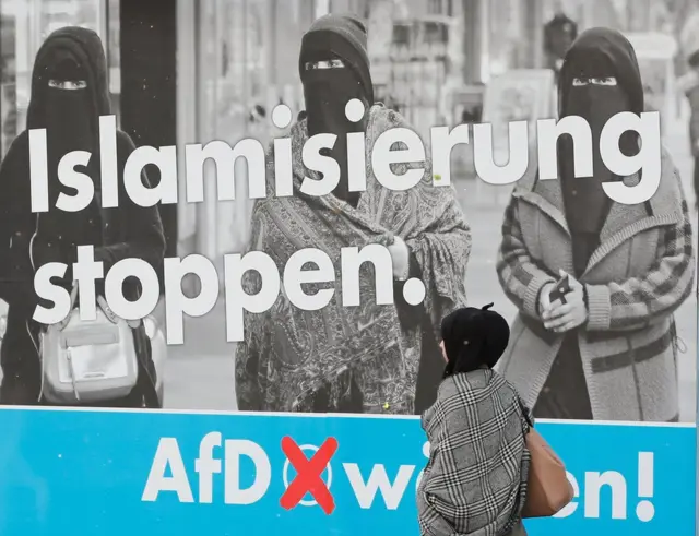 AfD'nin "İslamlaşmayı durdurun" yazan afişinin önünde yürüyen başörtülü bir kadın