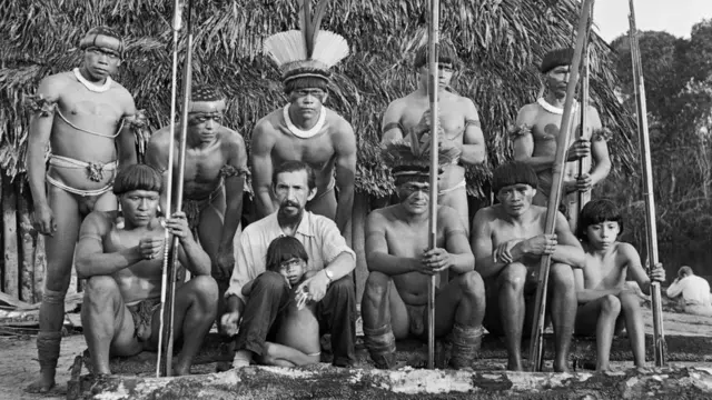 Fotografiacomo sacar o bonus da arbetypreto e branco mostra 9 homens do povo Yawalapiti, um homem branco e uma criança; parte do grupo está sentada