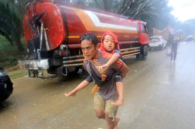 Seorang warga korban banjir mengendong anaknya untuk dievakuasi ke tempat keluarganya melintasi jalan lintas Nasional Medan-Banda Aceh yang terendam banjir di Desa Sungai Liput, Kecamatan Kejuruan, Aceh Tamiang, Aceh, Selasa (2/12/2025).