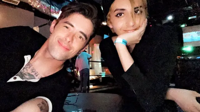 Eden with her friend Hayden. Both are smiling for the camera. Hayden has dark hair and tattoos on his chest and upper arm and is wearing a black top. Eden has blonde hair in this photo and is wearing a black hat and top. 