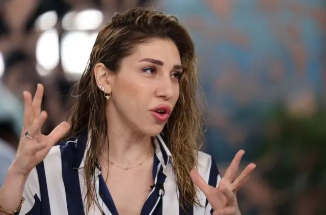 Kumral bir kadın, ellerini kullanarak konuşuyor. Üzerinde siyah beyaz bir gömlek var. 