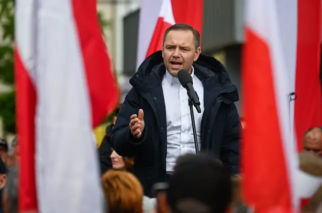 В центрі кадру – Кароль Навроцький перед мікрофоном із широко відкритим ротом і насупленими бровима. Він вдягнений у світло-блакитну сорочку, темну куртку та активно жестикулює. На передньому плані розфокусовані біло-червоні польські прапори та голови глядачів