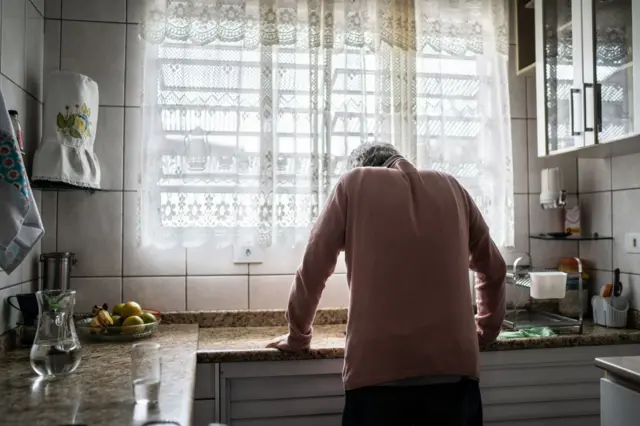 Um homem mais velho, de costas, apoiado na pia da cozinha, aparentemente exausto.