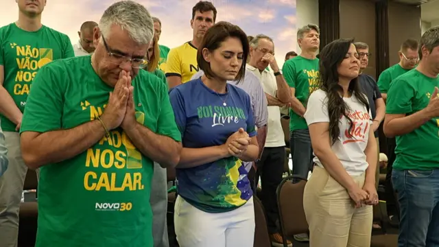 Michelle Bolsonaro veste uma camiseta azul onde se lê "Bolsonaro livre", ao lado do senador Eduardo Girão, que veste uma camiseta verde com os dizeres "Não vão nos calar", e da vereadora de Fortaleza Pricilla Costa, que veste uma camiseta branca escrita "Anistia Já". Ele têm os olhos fechados e estão em posição de oração, cercados de apoiadores bolsonaristas