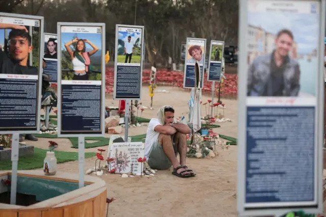 человек в белой футболке сидит на земле, вокруг него стоят стенды с фотографиями погибших на фестивале Supernova, у их подножья стоят цветы