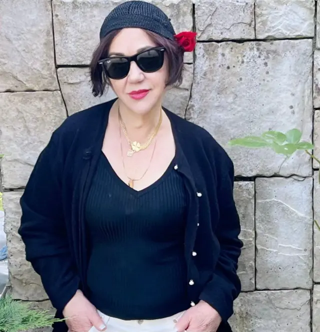 Öncel bir duvar önünde güneş gözlükleri beresi ve siyah üstüyle duruyor. 