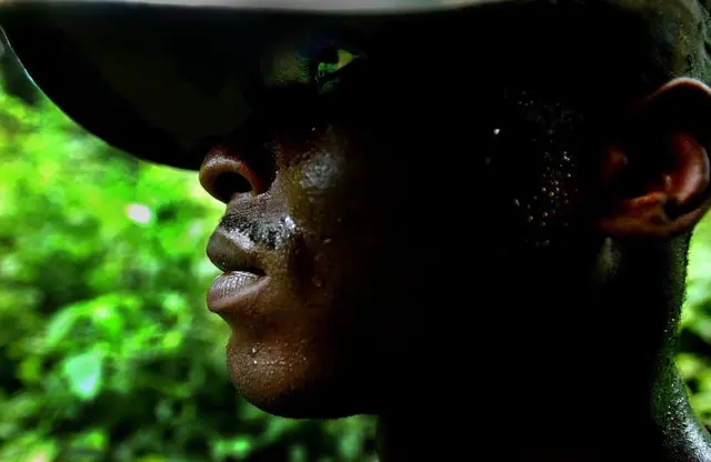 Un homme portant un chapeau, regarde ailleurs. Des herbes vertes sont visibles en arrière-plan.