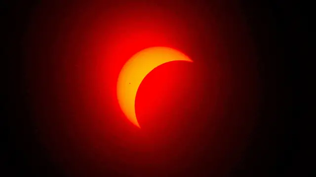 Foto colorida mostra processo do eclipse, com o Sol na partefoguetinho da betanotrás, laranja, e a lua o encobrindo como uma sombra