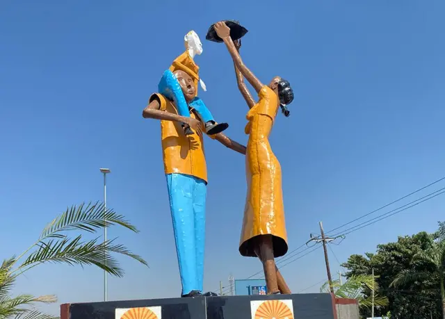 
Vista de baixo de uma estátua mostrando uma mulher entregando algo a uma criança que está sobre os ombros de um homem.
