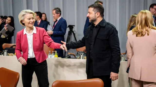 Глава Еврокомиссии Урсула фон дер Ляйен в красном пиджаке, темных брюках и белой рубашке протягивает руку Владимиру Зеленскому в темной одежде военного покроя. Зеленский тоже протягивает руку фон дер Ляйен