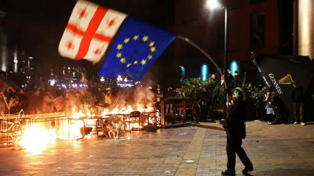 Протестующий размахивает флагами Грузии и ЕС перед горящей баррикадой во время митинга оппозиции в день местных выборов в Тбилиси.