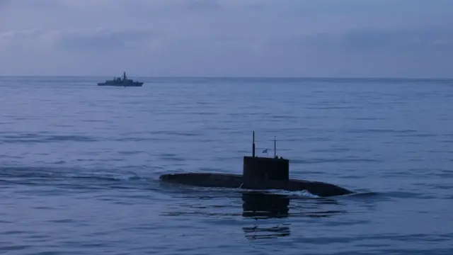 Подводная лодка в надводном положении в море, на заднем плане - военный корабль