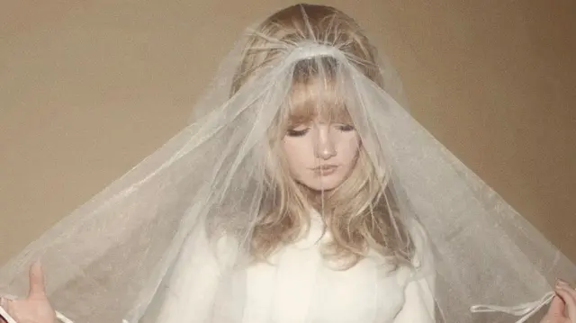 Una mujer de cabello rubio que lleva un vestido blanco y un velo de novia mira hacia el suelo.