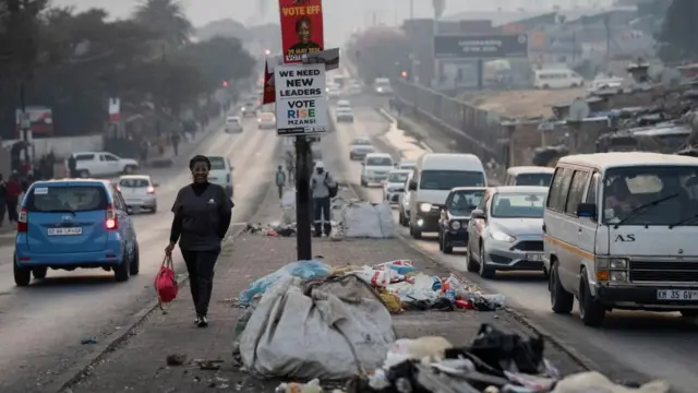 Umugore uhagaze ku muhanda iruhande rw'icyapa cyamamaza ishyaka EFF rya Julius Malema