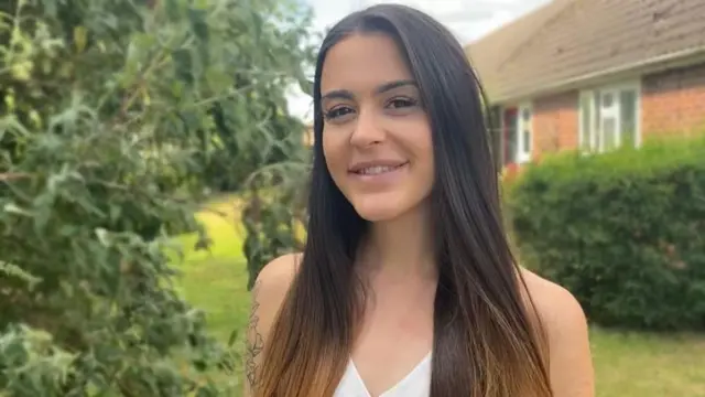 Abbie é uma mulher branca com top branco posando em frente a uma casa