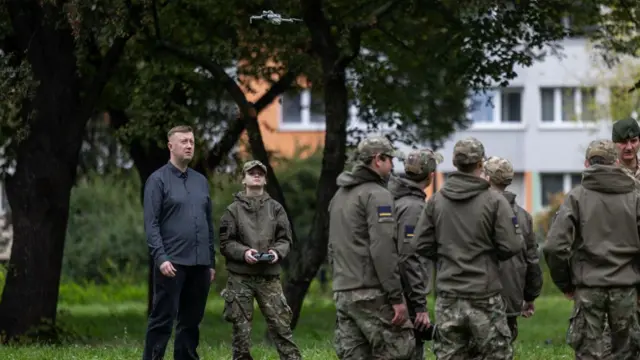 Kilku uczniów w mundurach wojskowych stoi na terenie szkoły. Nauczyciel w niebieskim ubraniu udziela im instruktażu. Jeden z uczniów lata dronem, który unosi się nad grupą.