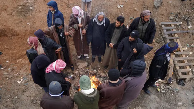 تجمع لأقارب سجناء حول نار قرب سجن الأقطان، حيث يُحتجز بعض معتقلي تنظيم الدولة، 22 يناير/كانون الثاني 2026، وجميعهم رجال يرتدي بعضهم جلاليب والبعض الآخر يرتدي سراويل وسترات، بينما يغطي بعض منهم رأسه بشيلان مصنوعة من القماش