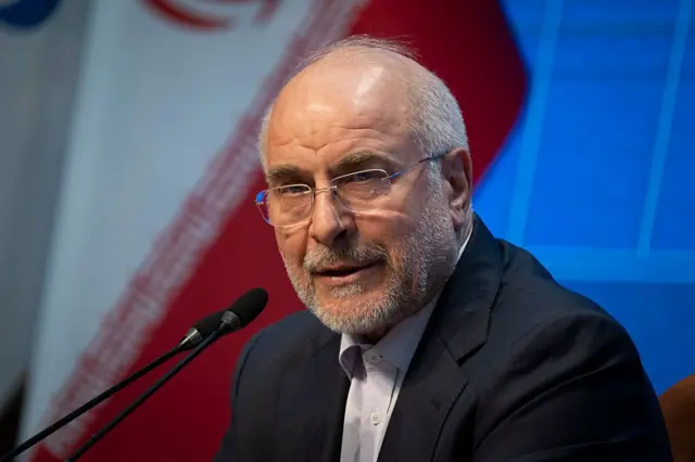 محمدباقر قالیباف پشت میکروفون در پسزمینه رنگ آبی و پرچم جمهوری اسلامی دیده می‌شود