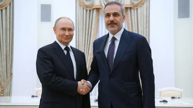 Hakan Fidan, Kremlin'de Vladimir Putin ile el sıkışıyor ve kameralara bakıyorlar. İkisi de tebessüm ediyor