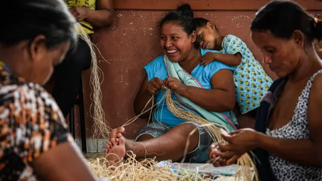 Três mulheres warao fazem artesanato, uma delas tem um grande sorriso e uma criança apoiada no ombro