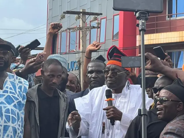 Issa Tchiroma vêtu d’un grand boubou blanc traditionnel et coiffé d’un bonnet rouge et noir orné, tient un micro et s’adresse à la foule. Plusieurs hommes l’entourent de près.