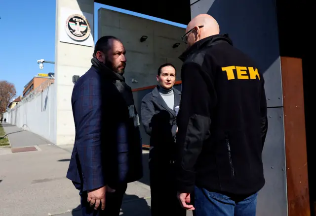 Троє людей стоять біля входу до масивної будівлі з бетону та металу, яку позначає емблема на стіні. Одна людина одягнена в темну куртку з великим написом "TEK" на спині, а двоє інших стоять навпроти, ведучи розмову. Місце виглядає як охоронюваний об’єкт із високим парканом і воротами вздовж вулиці.