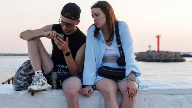 Юноша и девушка сидят на парапете набережной и смотрят в телефон