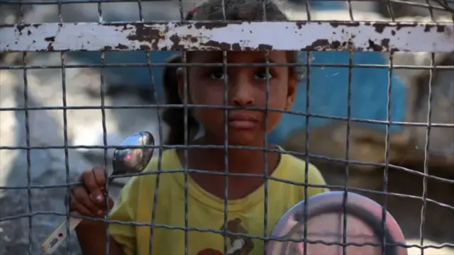 تصویر یک کودک گرسنه فلسطینی که یک قاشق و بشقاب خالی را بدست دارد و از پشت یک پنجره سیمی نگاه می‌کند.