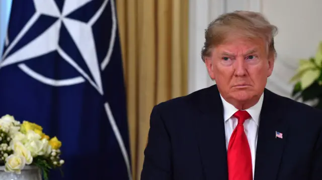 Donald Trump podczas szczytu NATO w czasie swojej pierwszej kadencji jako prezydent, stojący na tle flagi NATO.