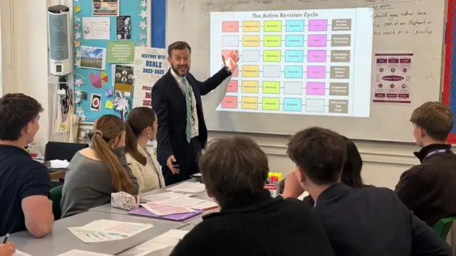 A man wearing a suit pointing at a whiteboard with revision information to a group of students wearing black blazers. 