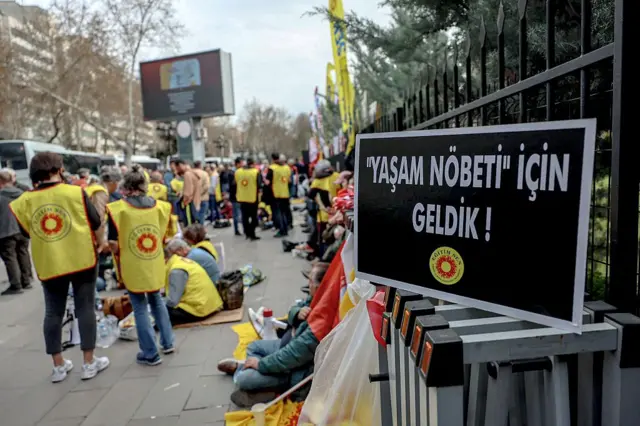 Fotoğrafta 15 Nisan 2026 tarihinde Ankara'daki Milli Eğitim Bakanlığı binası önünde, okul saldırılarını protesto etmek amacıyla eğitim sendikaları üyelerinin düzenlediği oturma eylemi sırasında, üzerinde “Yaşam nöbeti için geldik” yazan bir pankart görülüyor. 
