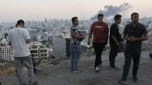 Wanaume kadhaa wamesimama juu ya mlima wakitazama moshi ukitoka kwenye jengo la shirika la utangazaji la taifa la Iran baada ya kulipuliwa na Israel.