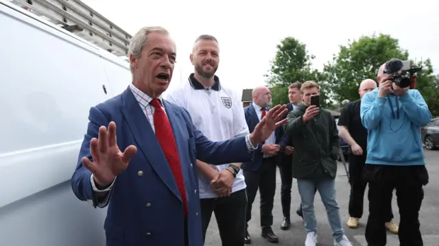 Nigel Farage, lider partii Reform UK, przemawia z uniesionymi rękami, stojąc obok białego samochodu dostawczego. Za nim stoi lekko uśmiechnięty kandydat partii na deputowanego z okręgu Makerfield, Robert Kenyon. Za nimi, po prawej stronie zdjęcia, stoi grupa mężczyzn. Jeden z nich trzyma w górze kamerę wideo z dołączonym mikrofonem. Inny trzyma w górze smartfon. 