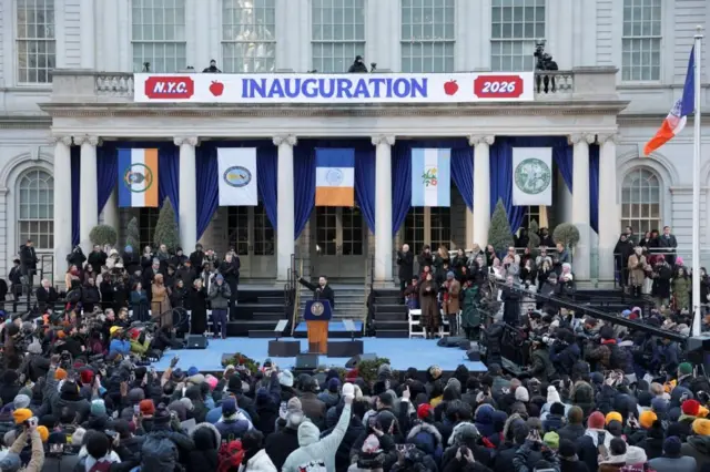 市役所前の舞台に登壇し演説しながら右手を上げるマムダニ新市長と、周りでそれを見守る大勢の人たち