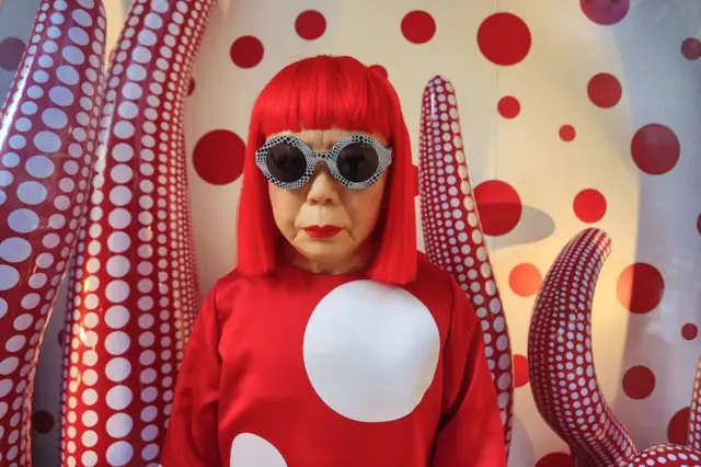 yayoi kusama 2012 yılında üzerinde kendi imzası haline gelmiş puantiyeli bir sweatshirtle ve güneş gözlükleriyle poz veriyor.