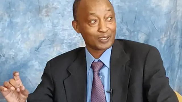 Dr Théogène Rudasingwa akoze ikimenyetso n'ikiganza yicaye mu ntebe. Yambaye ikositimu y'umukara, karuvati y'iroza n'ishati y'ubururu. Ku ikoti rye hometseho indangururamajwi ntoya. Arimo kuvuga areba ku ruhande.