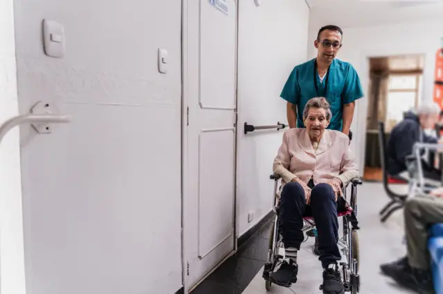 Senhora sendo empurrada em cadeira de rodas por profissional, com outros idosos ao fundo