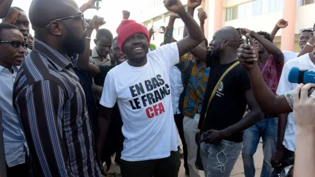 Plusieurs hommes applaudissent à l'extérieur. L'homme au centre porte un bonnet rouge et un t-shirt blanc.