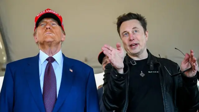 Donald Trump usando um boné vermelho com a inscrição Make America Great Again ao lado de Elon Musk, que está falando e gesticulando.
