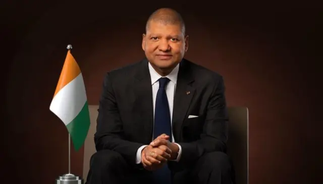 Jean Louis Billon, en costume sombre, cravatte bleue, avec le drapeau ivoirien posé à sa droite