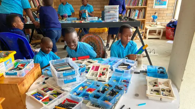 Abana benshi bari mu ishuri bafite ibikoresho bitandukanye biri ku meza. Umwe muri bo arimo guseka, undi arimo kumwenyura. Bambaye imyambaro y'ishuri y'ubururu. 