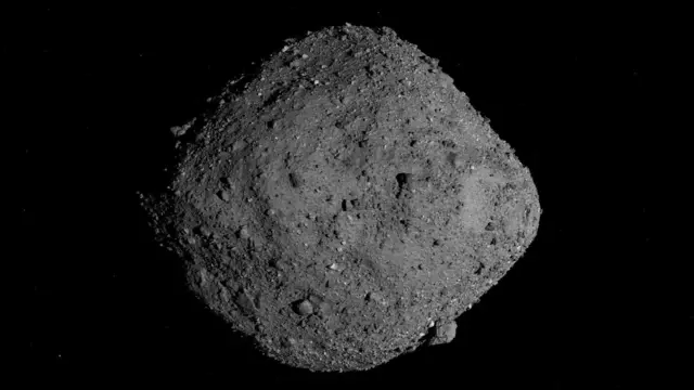 Imagem de um asteroide de 500 m de largura chamado Bennu que parece uma rocha cinza que é mais larga no meio do que em cada extremidade. O objeto não é liso — há protuberâncias nodosas de tamanhos diferentes saindo de sua superfície.